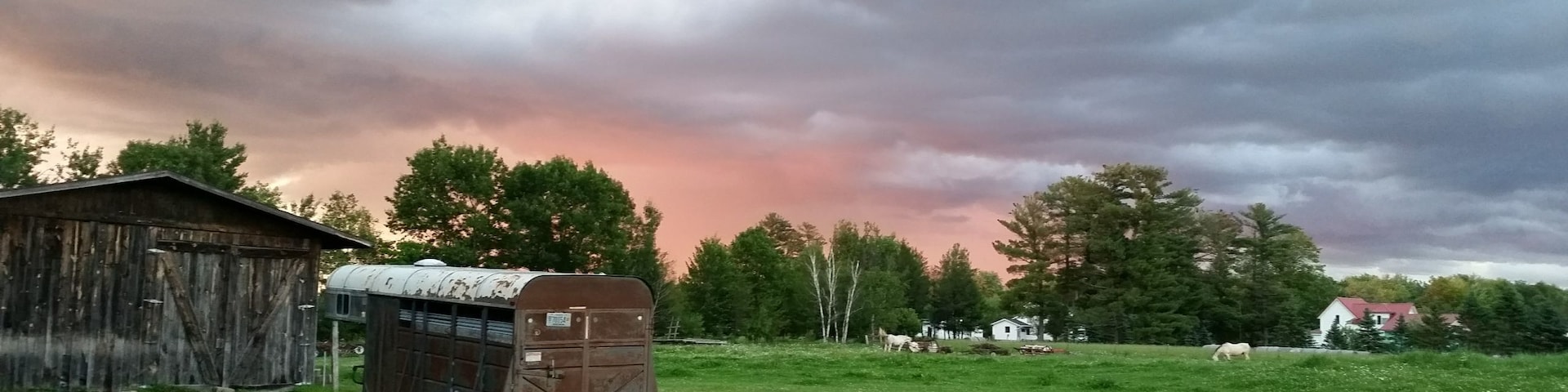 Paisagem rural com uma casa rodante, um celeiro e cavalos pastando sob um céu nublado.