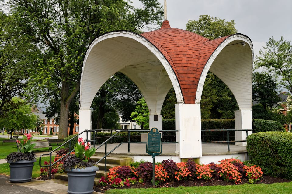 Ein Gazebo mit rotem Dach und weißen Wänden, umgeben von Blumenbeeten und Bäumen.