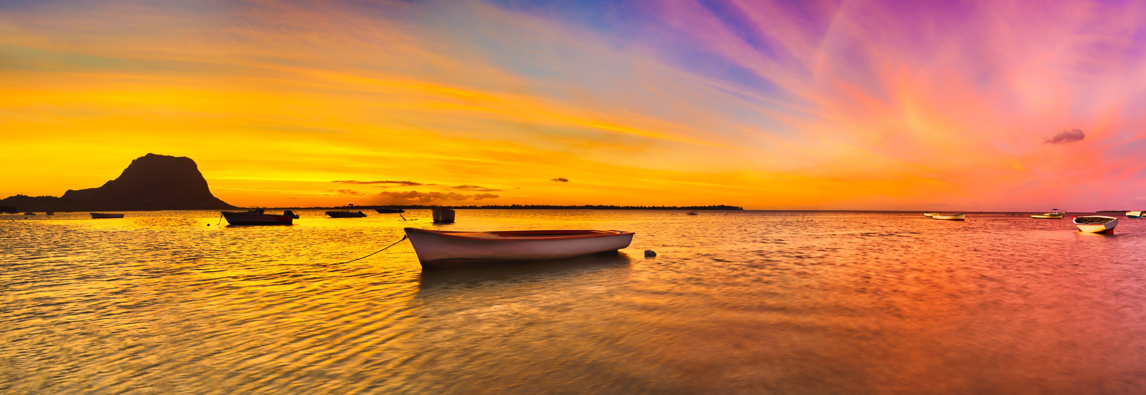 A sunset over a calm sea with boats and a prominent mountain silhouette.