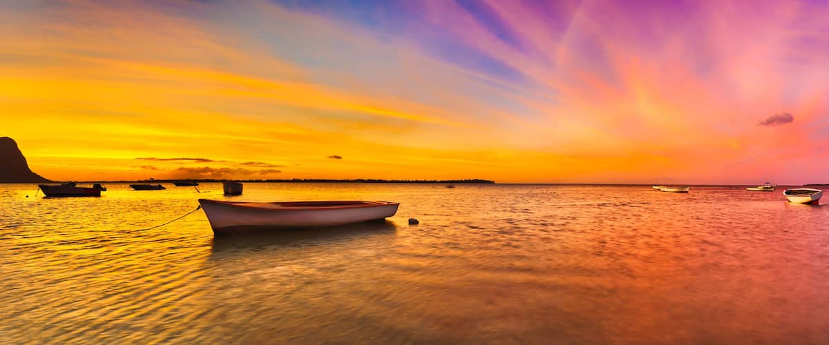 A sunset over a calm sea with boats and a prominent mountain silhouette.