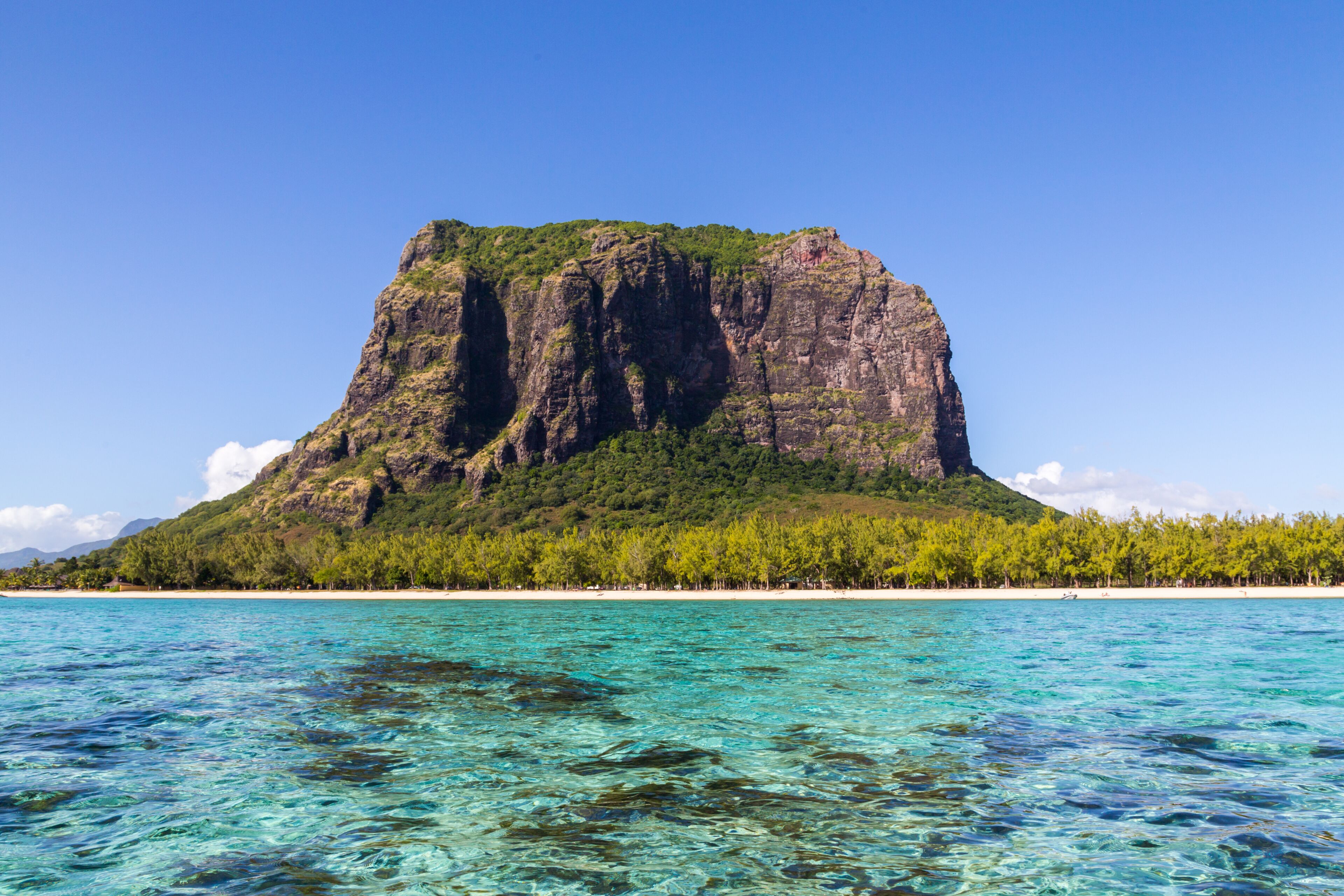 Um mar azul-turquesa transparente, uma ilha rochosa com vegetação verde exuberante e uma linha de costa distante.