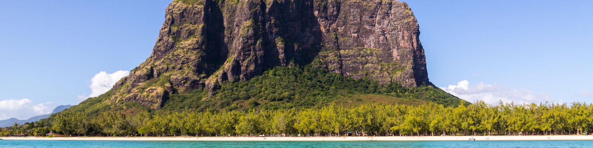Um mar azul-turquesa transparente, uma ilha rochosa com vegetação verde exuberante e uma linha de costa distante.