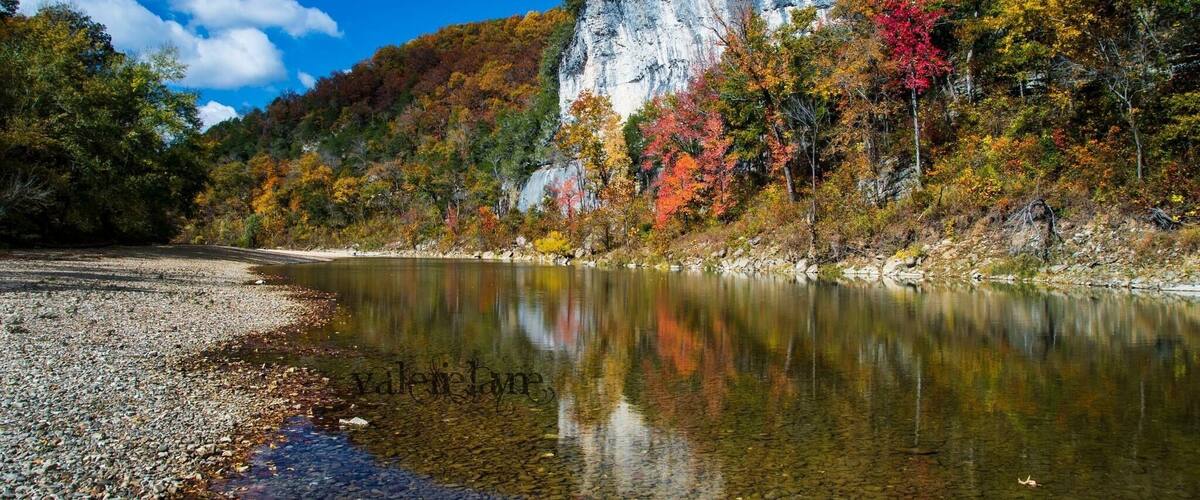 A calm river with clear water reflecting the surrounding autumn foliage and a rocky shoreline.