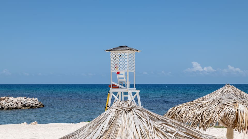 A beach with a lifeguard tower, thatched umbrellas, and a calm sea.