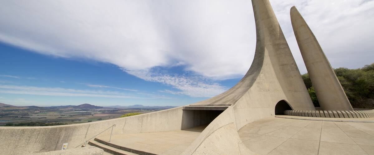 A concrete monument with three tall, curved structures against a backdrop of a clear sky and distant landscape.