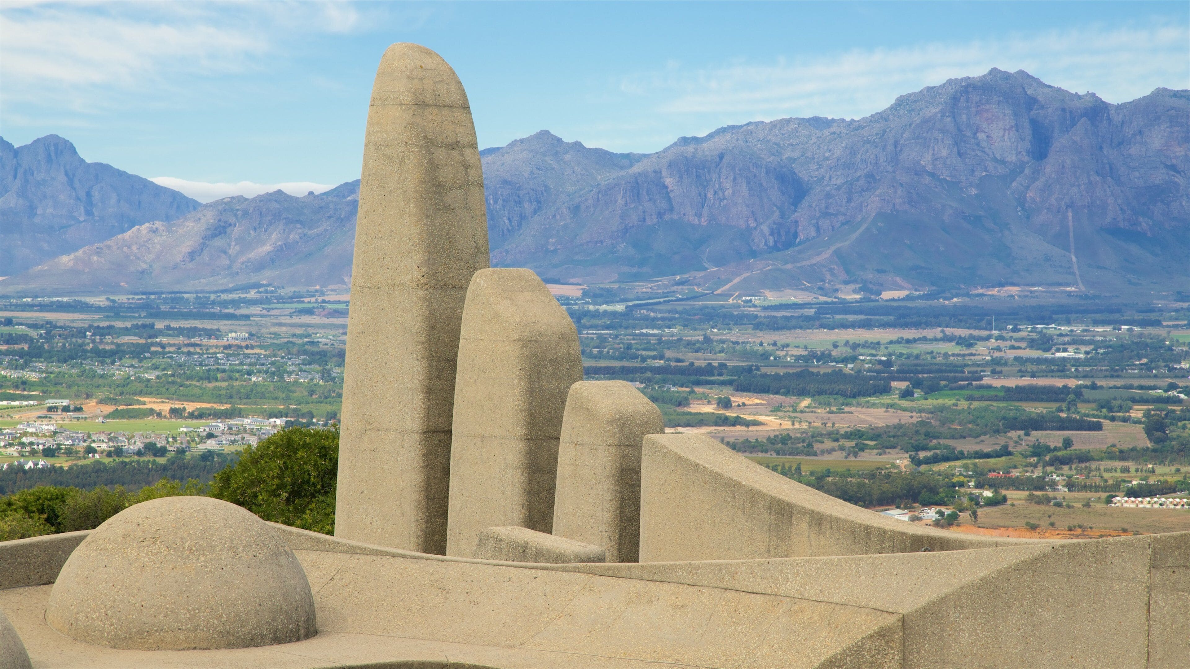 A large stone monument with a tall, narrow obelisk and two shorter, curved structures, set against a backdrop of mountains and a valley with scattered buildings.
