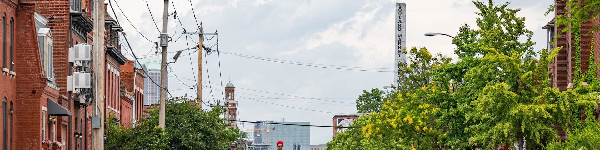 A street lined with parked cars and buildings, a cyclist in the foreground, and a large sign reading "SOULARD MARKET" in the background.