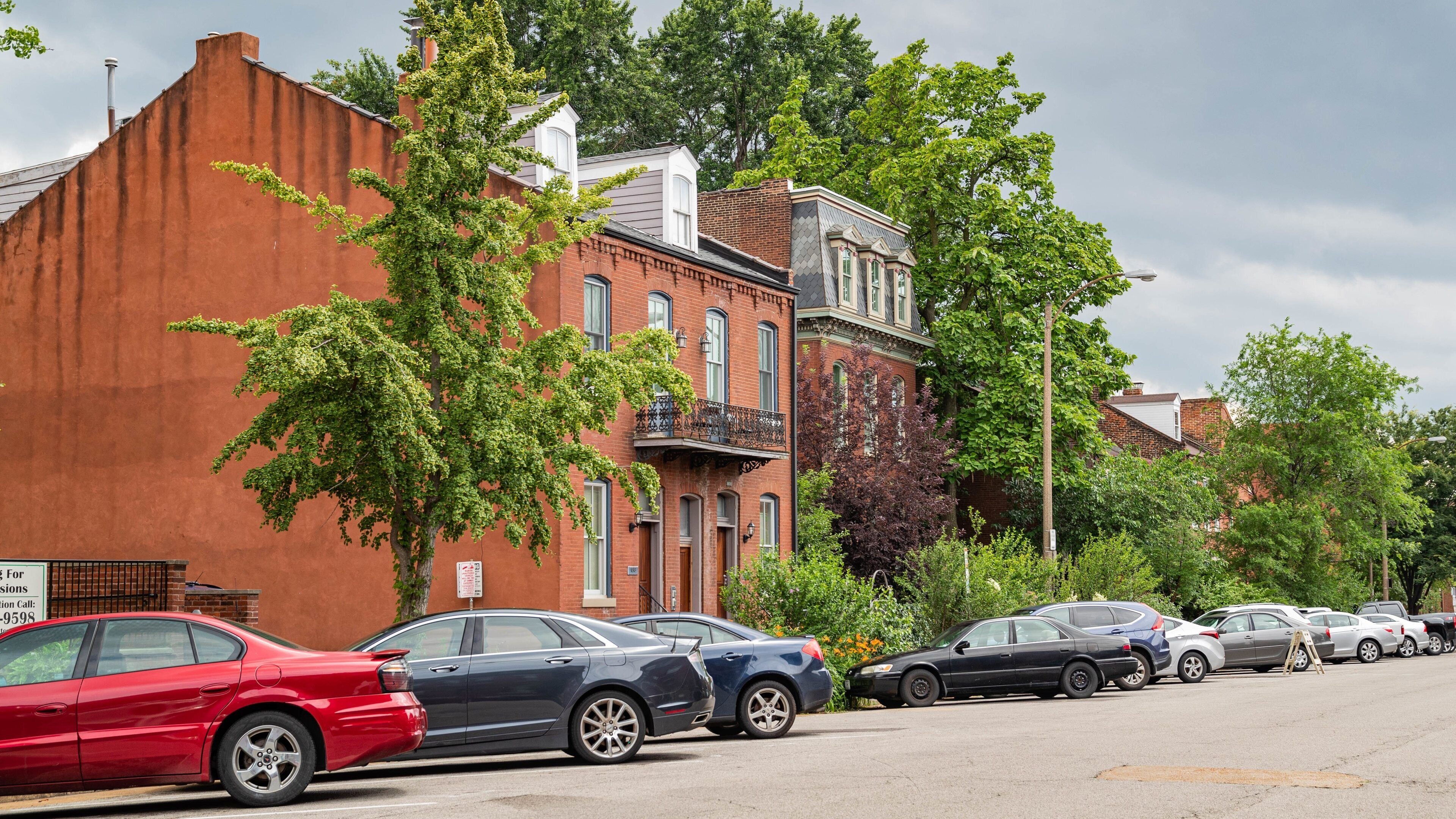 A row of parked cars along a tree-lined street with historic brick buildings.
