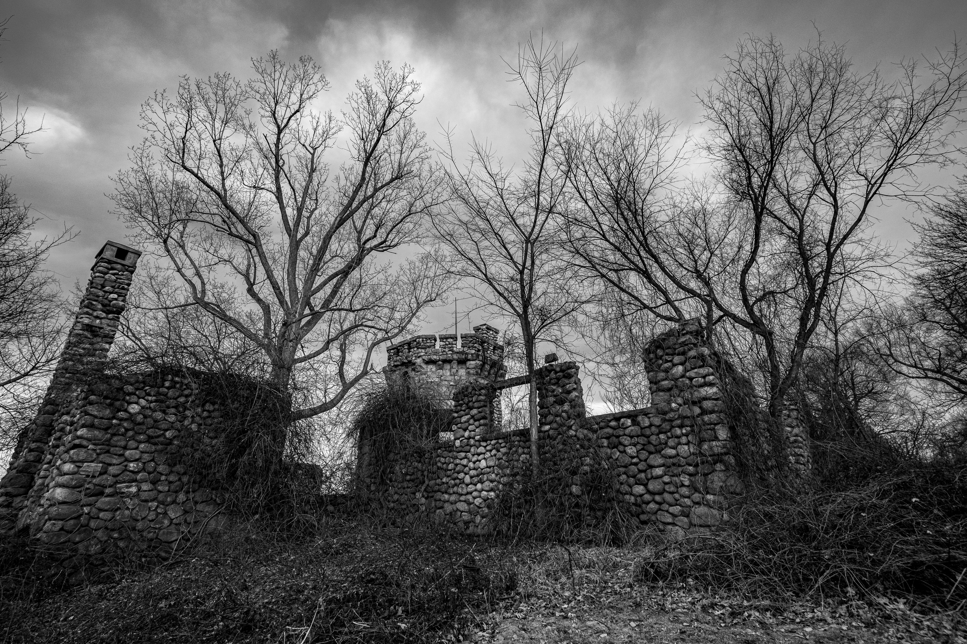 Une photographie en noir et blanc représentant les ruines d’une construction en pierre, avec des arbres dénudés en arrière-plan.