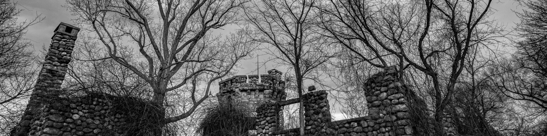 Une photographie en noir et blanc représentant les ruines d’une construction en pierre, avec des arbres dénudés en arrière-plan.
