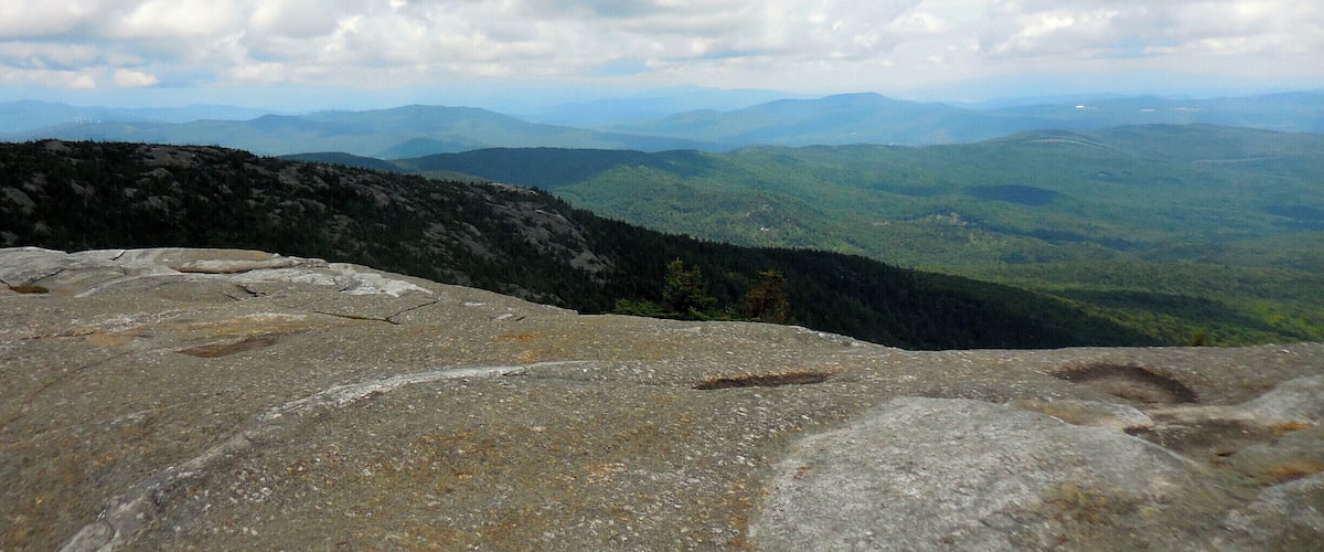 A mountainous landscape with rocky outcrops and a vast view of rolling hills under a cloudy sky.