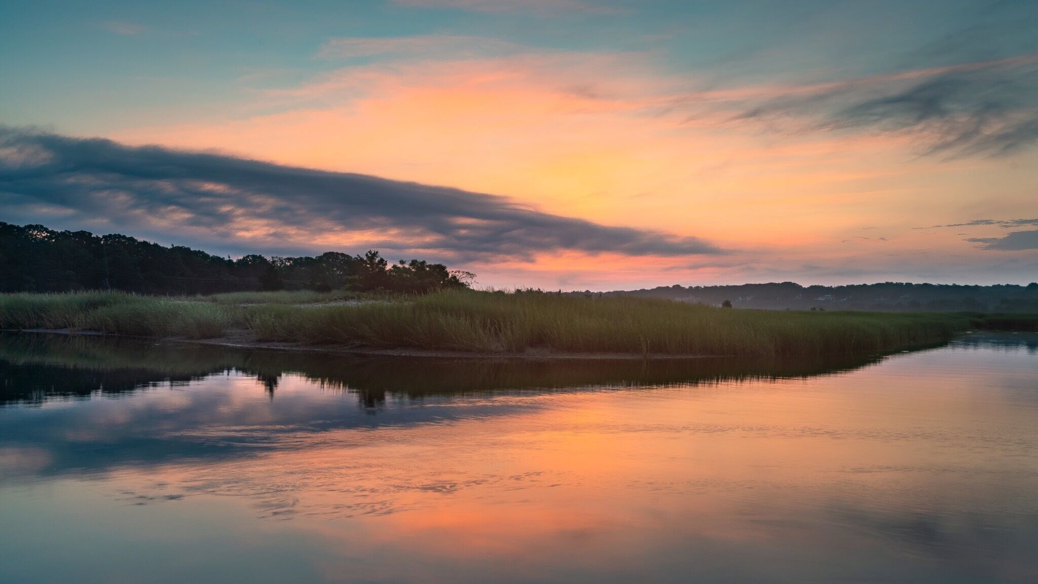 A calm river reflecting the sunset sky, surrounded by grassy marshland and distant trees.