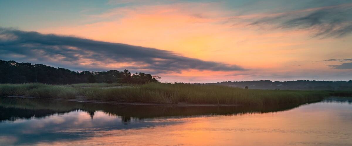 A calm river reflecting the sunset sky, surrounded by grassy marshland and distant trees.