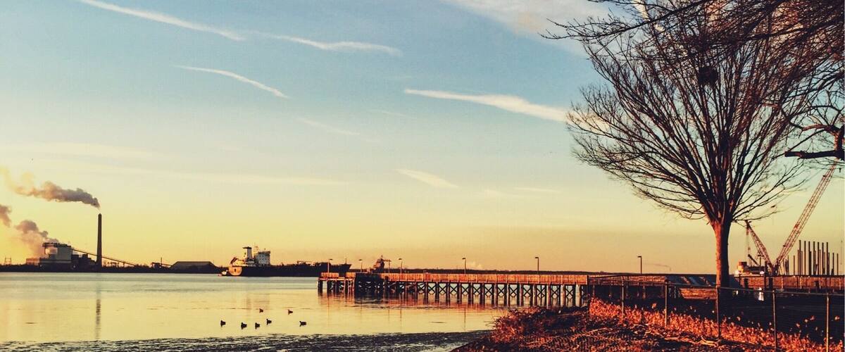 A waterfront scene with a pier extending into the water, a tree on the right, and industrial structures in the background.