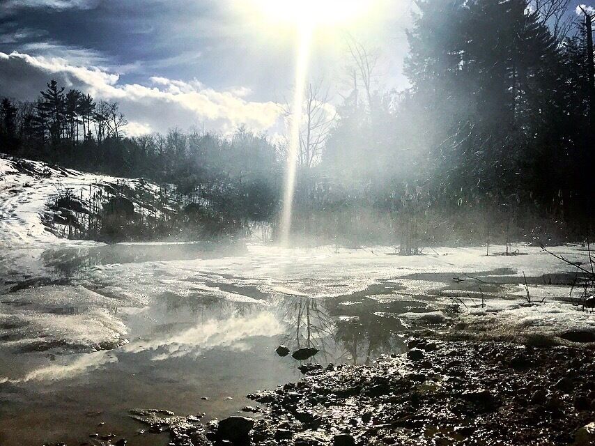 A frozen river with sunlight reflecting on the ice, surrounded by trees and a cloudy sky.