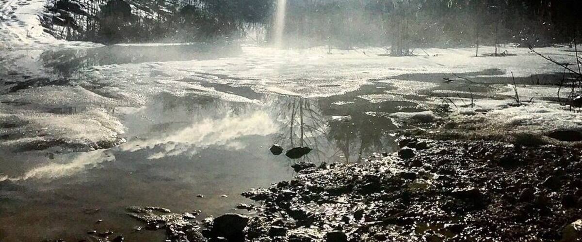 A frozen river with sunlight reflecting on the ice, surrounded by trees and a cloudy sky.