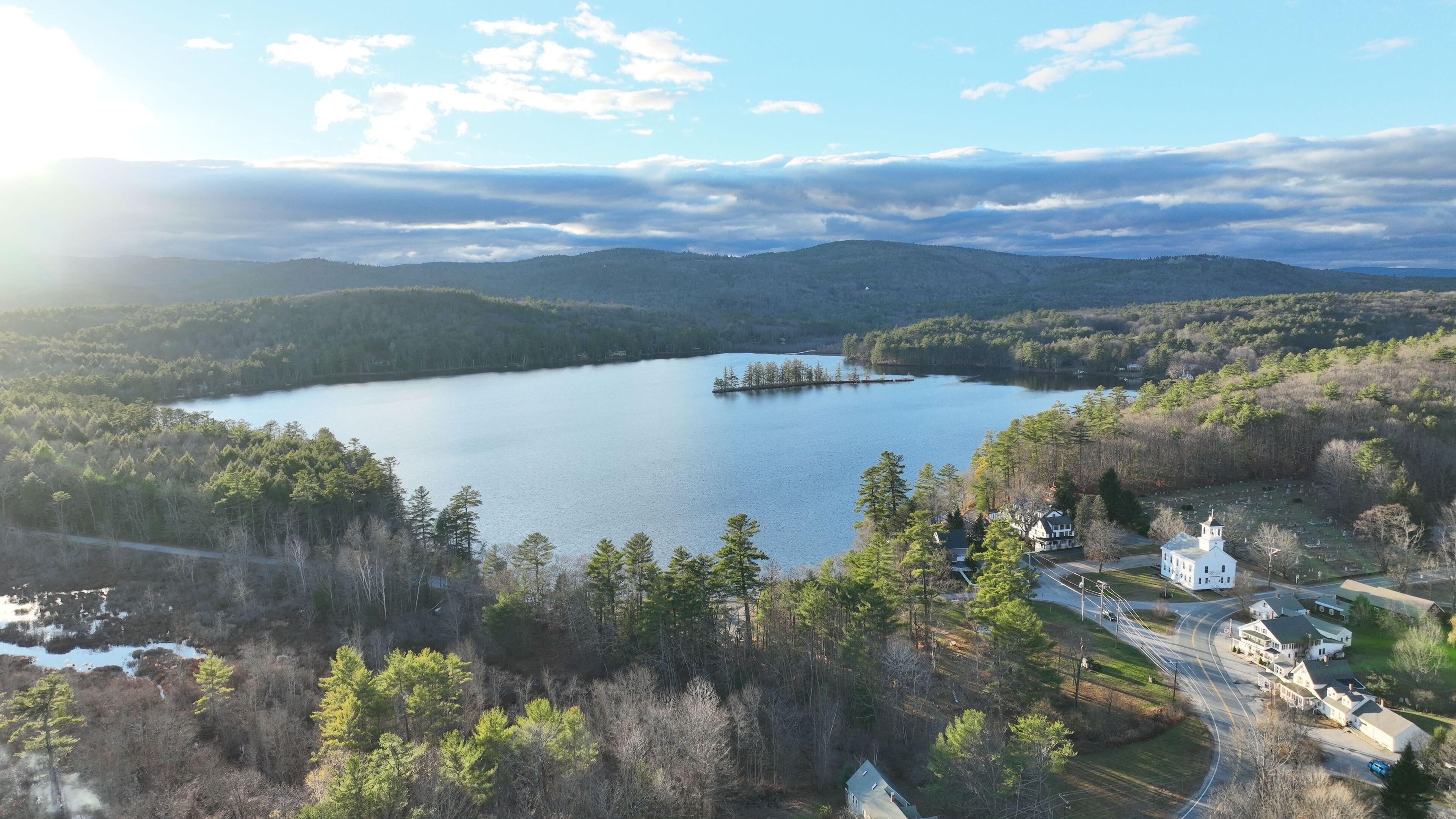 A lake surrounded by forested hills, a small island, and a cluster of white houses near a road.