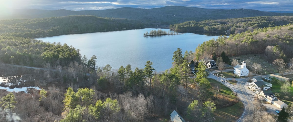 A lake surrounded by forested hills, a small island, and a cluster of white houses near a road.