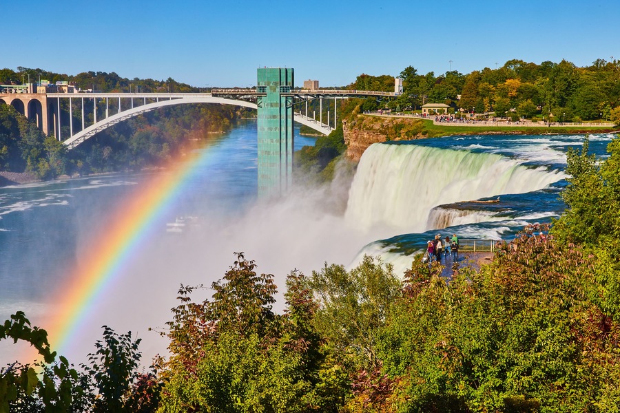 A waterfall with a rainbow, a bridge, and lush greenery.