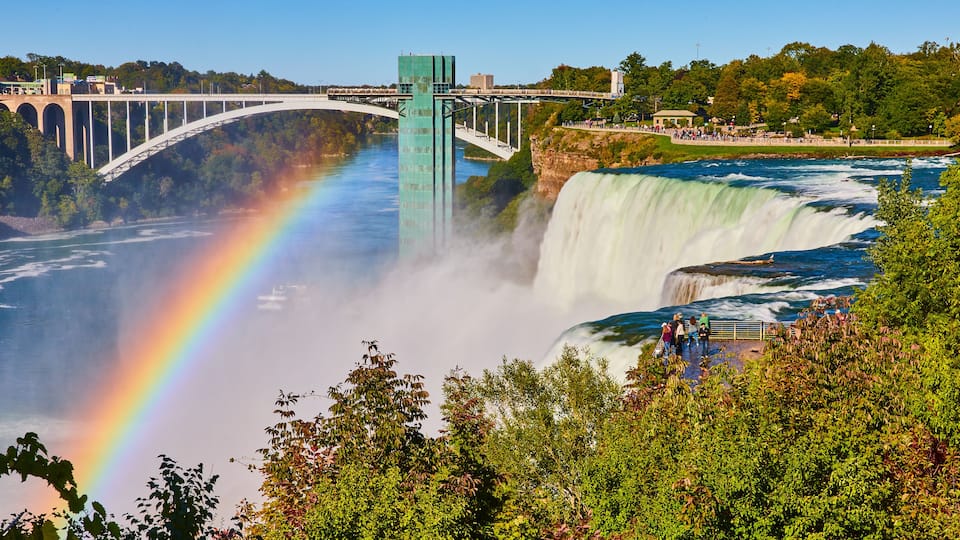 A waterfall with a rainbow, a bridge, and lush greenery.