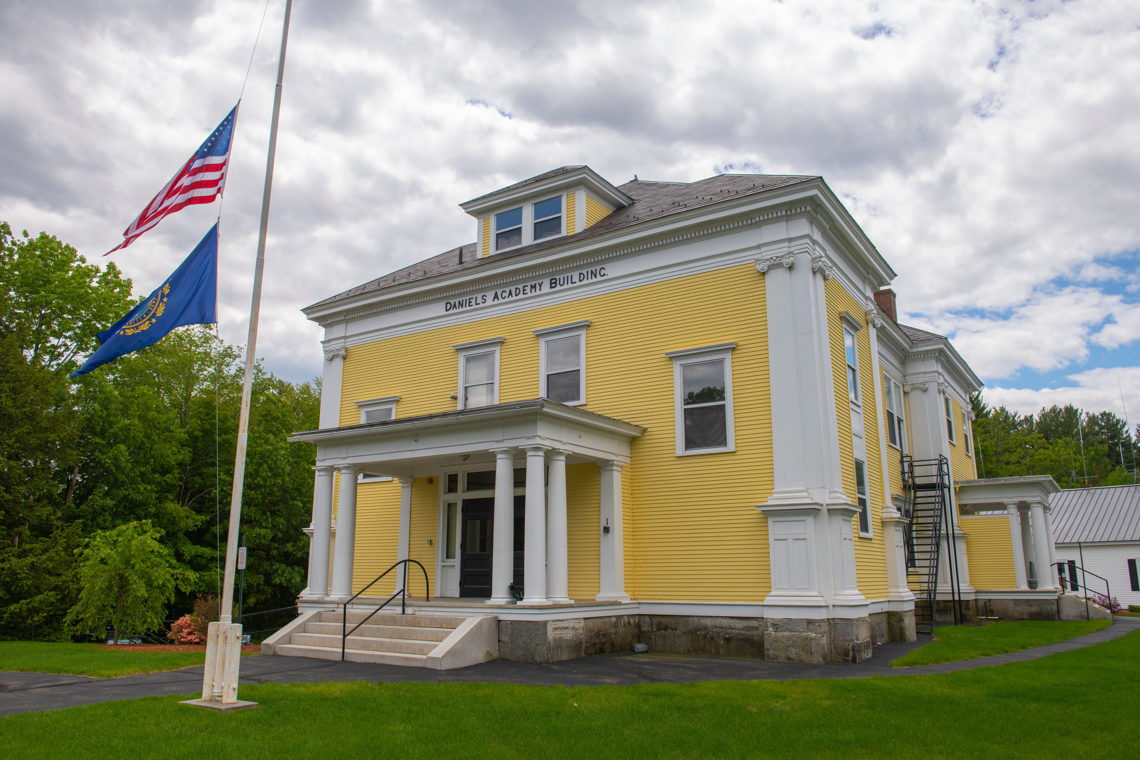 Um edifício amarelo com a inscrição 'DANIELS ACADEMY BUILDING' na fachada, um mastro com a bandeira americana e uma bandeira azul com desenhos dourados.