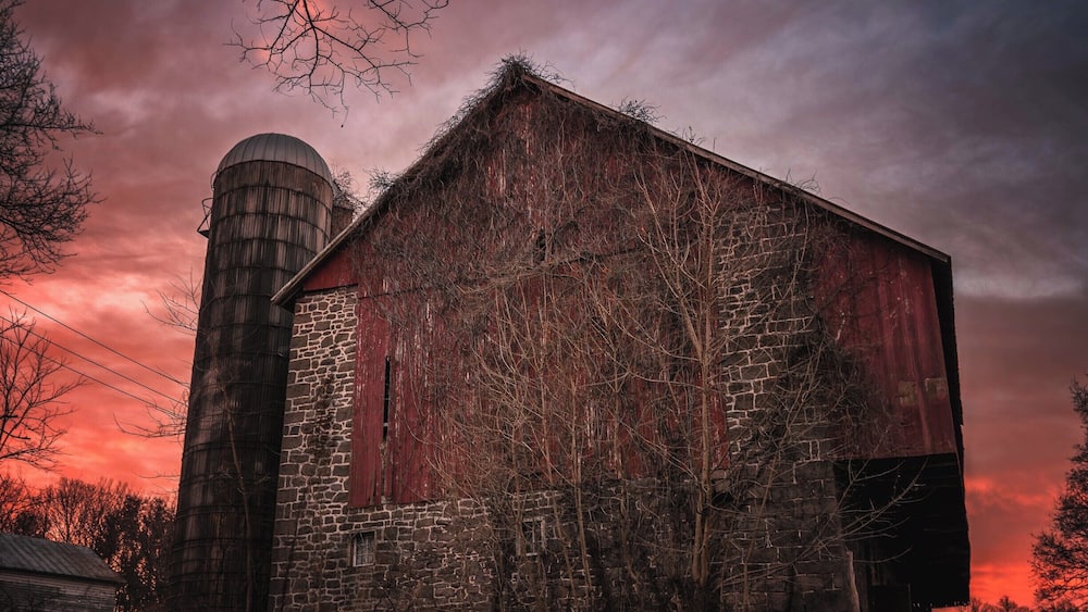 A rustic barn with a silo, surrounded by bare trees and a dramatic sunset sky.
