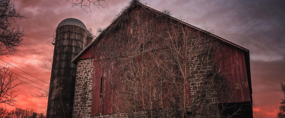 A rustic barn with a silo, surrounded by bare trees and a dramatic sunset sky.