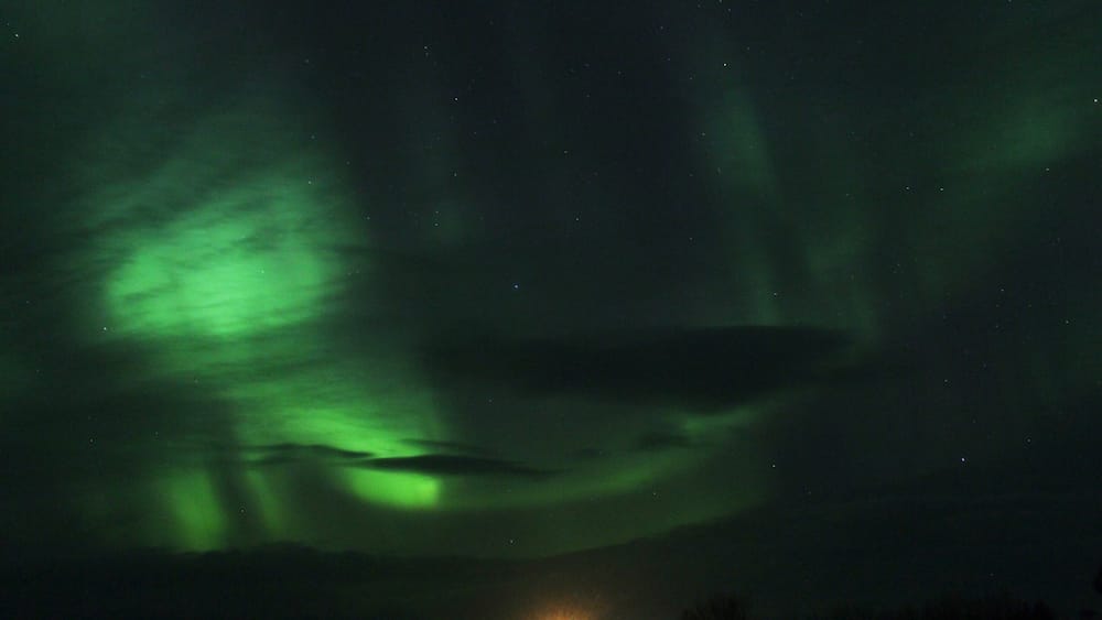 The Northern Lights over a dark sky with a faintly lit building at the bottom.