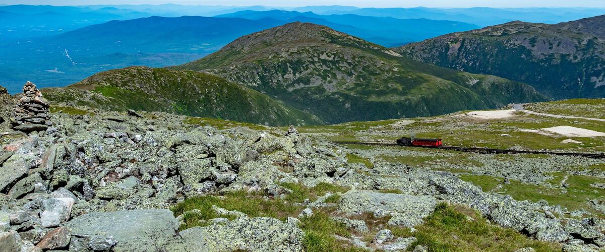 A mountain landscape with a train on a narrow track, rocky terrain, and distant mountains under a clear blue sky.