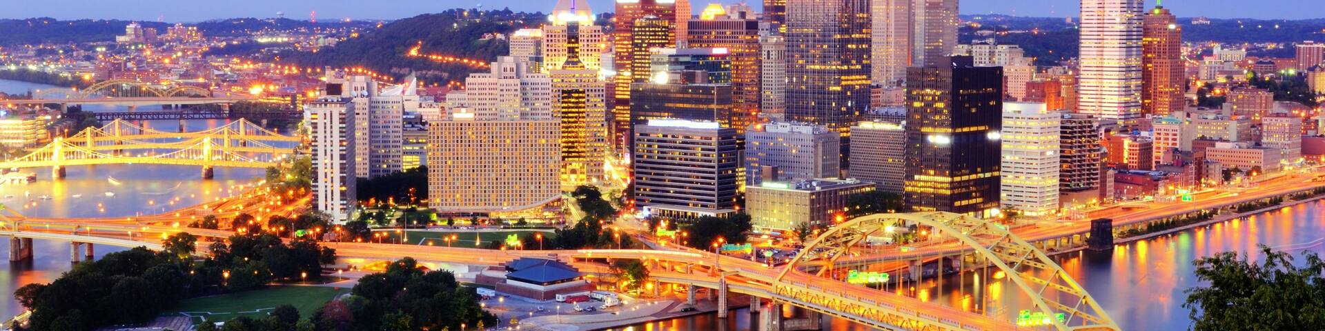 A city skyline at dusk with illuminated buildings, a river, and a bridge.
