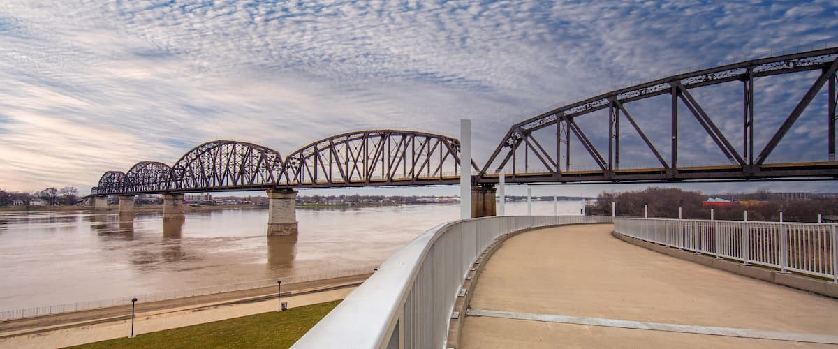 A steel bridge with multiple arches over a river, a concrete viewing platform, and a cloudy sky.