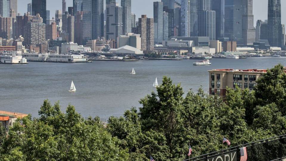A city skyline with prominent skyscrapers, a river with sailboats, and a sign reading 'WEST NEW YORK' with American flags.