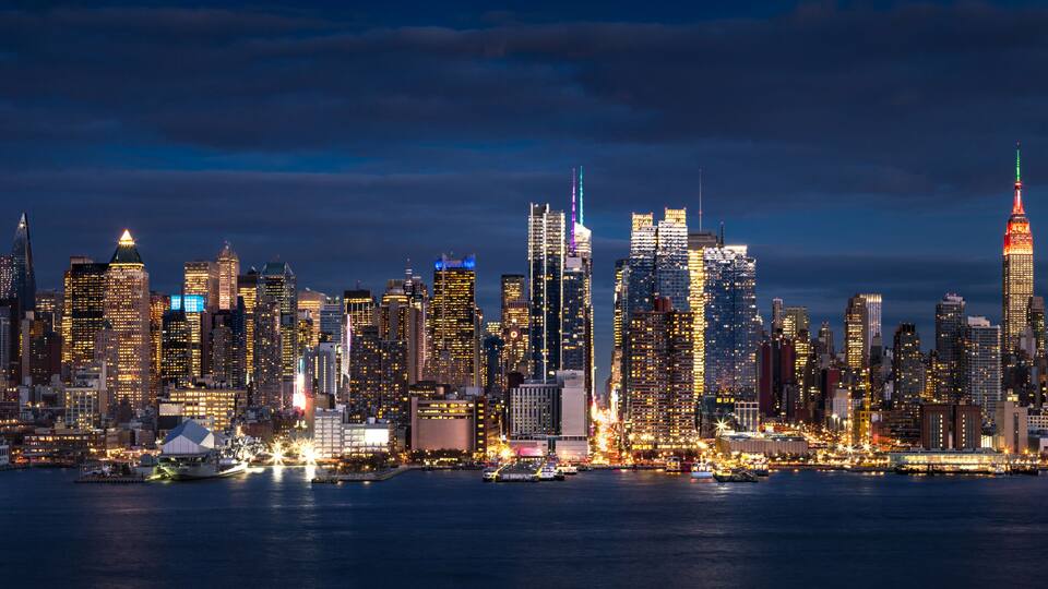 A city skyline at night with illuminated buildings and a body of water in the foreground.