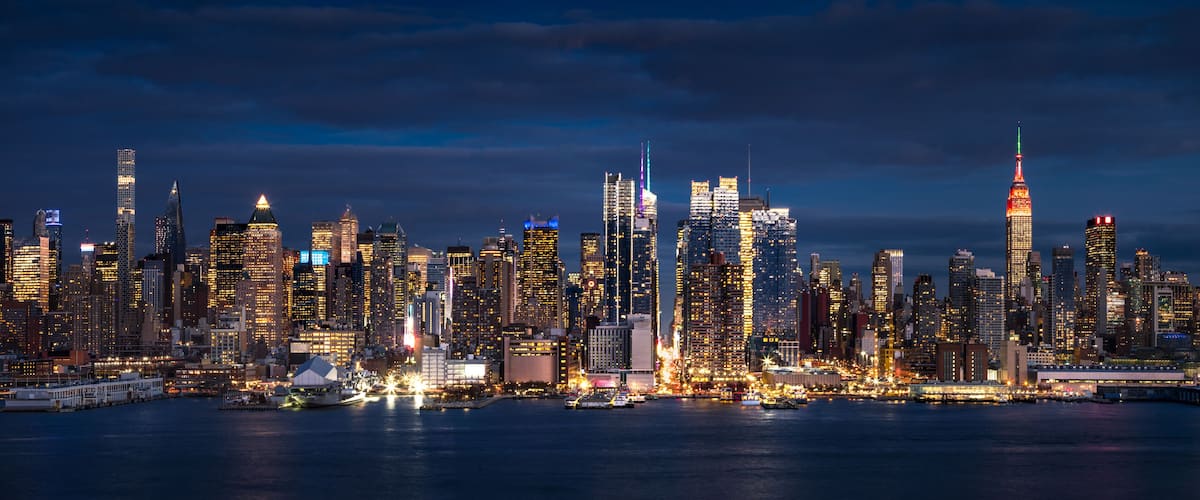 A city skyline at night with illuminated buildings and a body of water in the foreground.