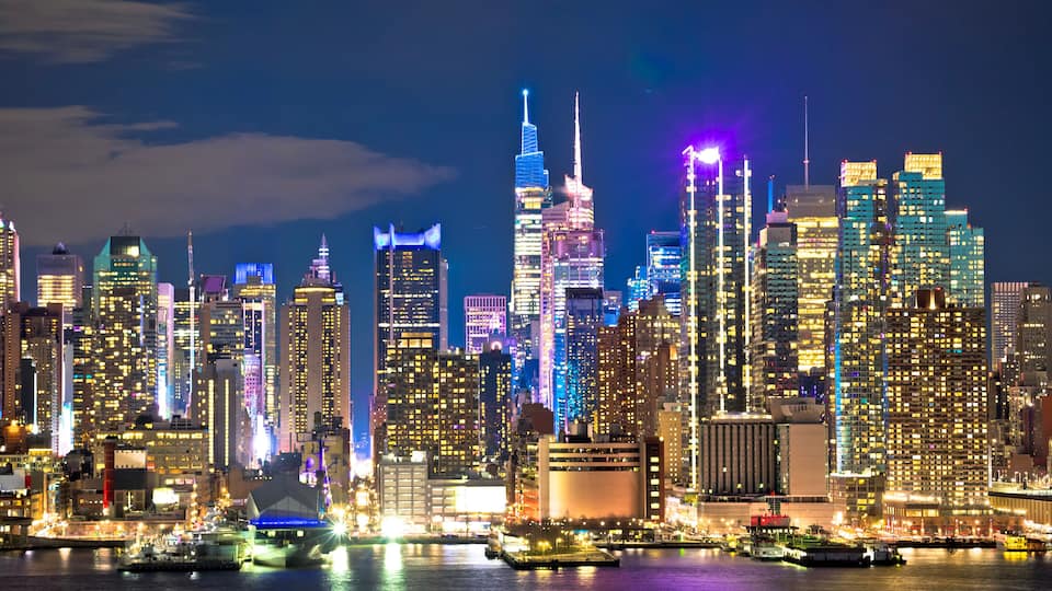 A city skyline with illuminated skyscrapers and a body of water in the foreground.