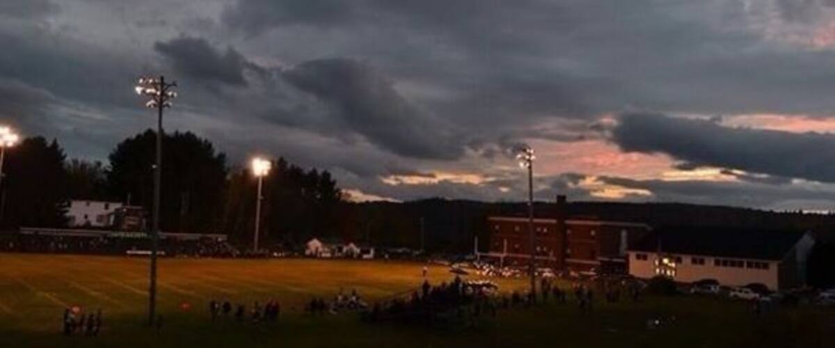 A sports field at dusk with floodlights, a building, and a cloudy sky.