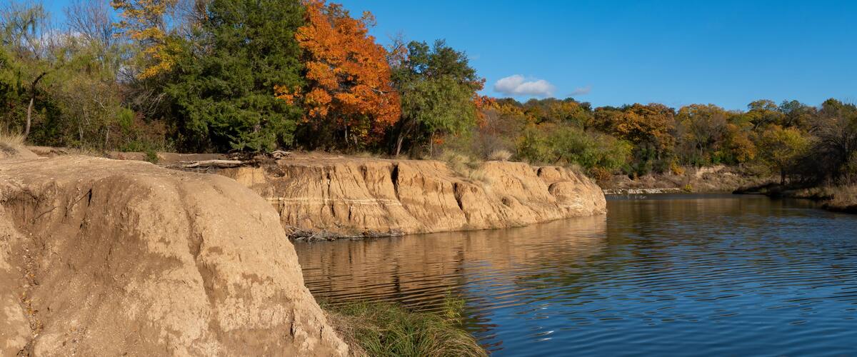 A riverbank with a concrete dam, autumn-colored trees, and a clear blue sky.