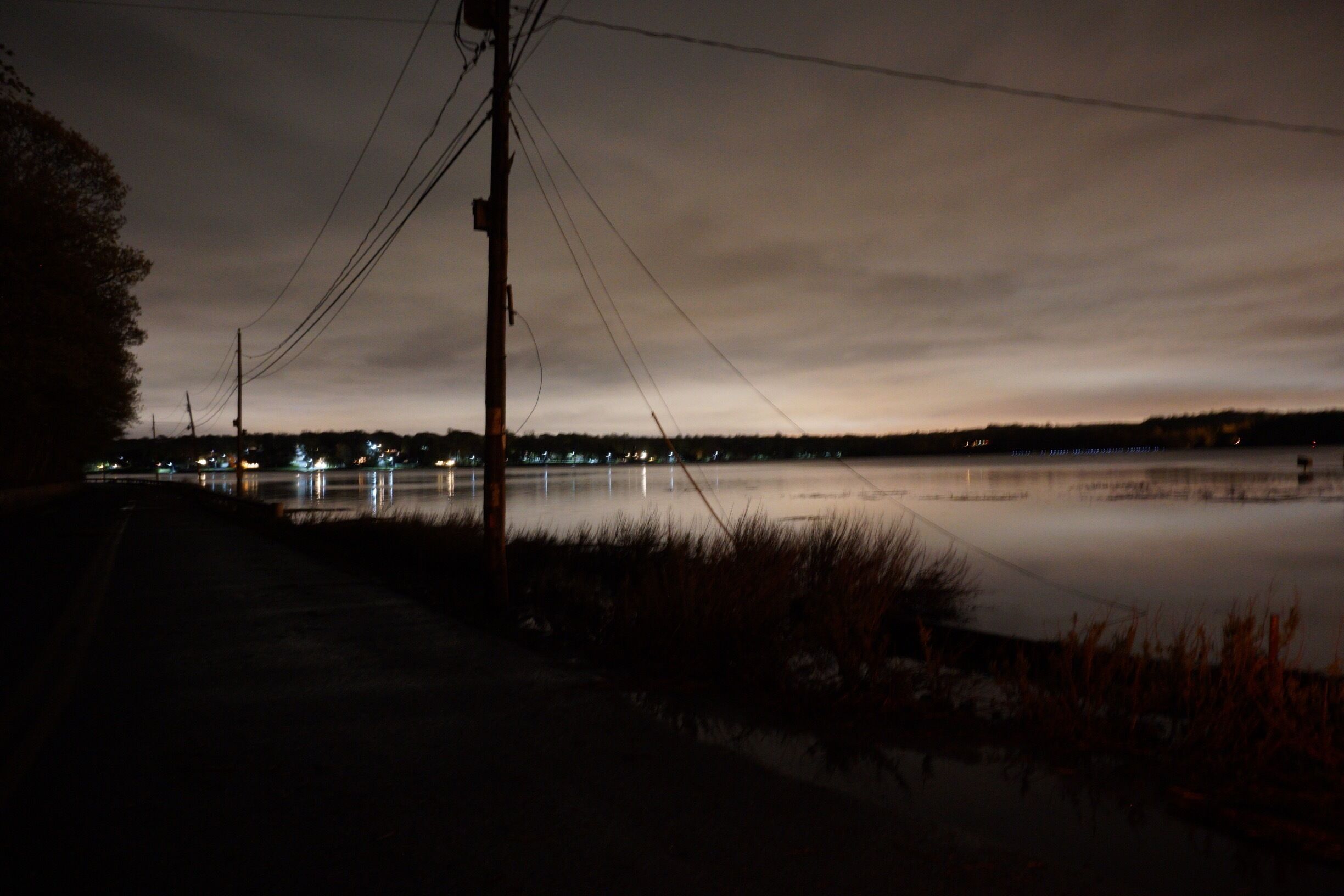 A nighttime view of a calm lake with a road and power lines in the foreground.