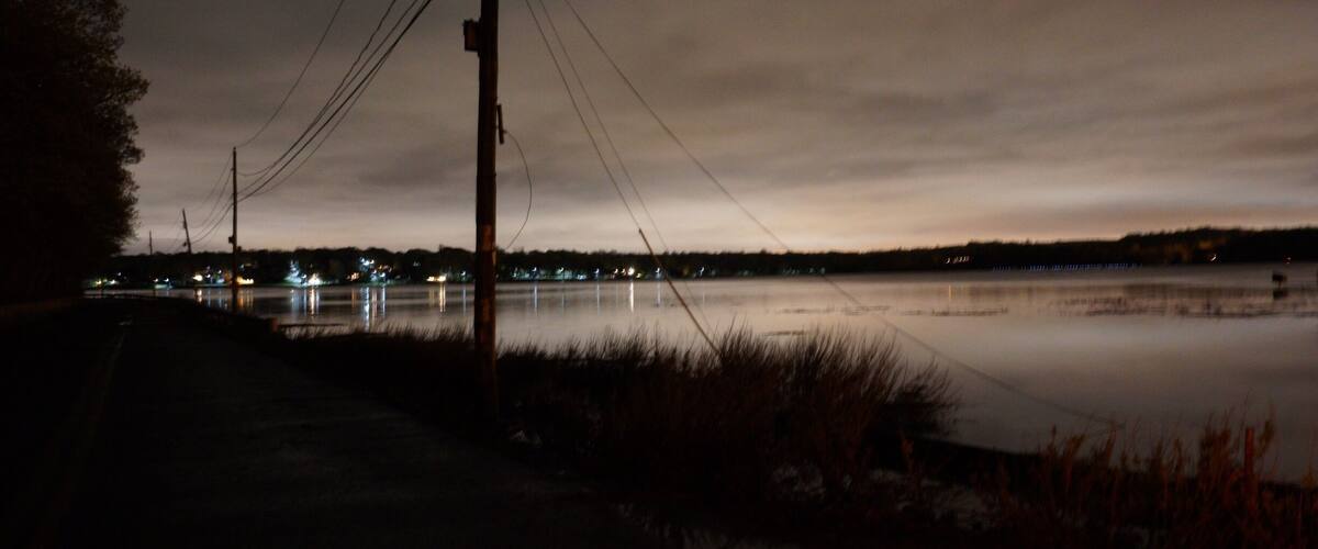 A nighttime view of a calm lake with a road and power lines in the foreground.