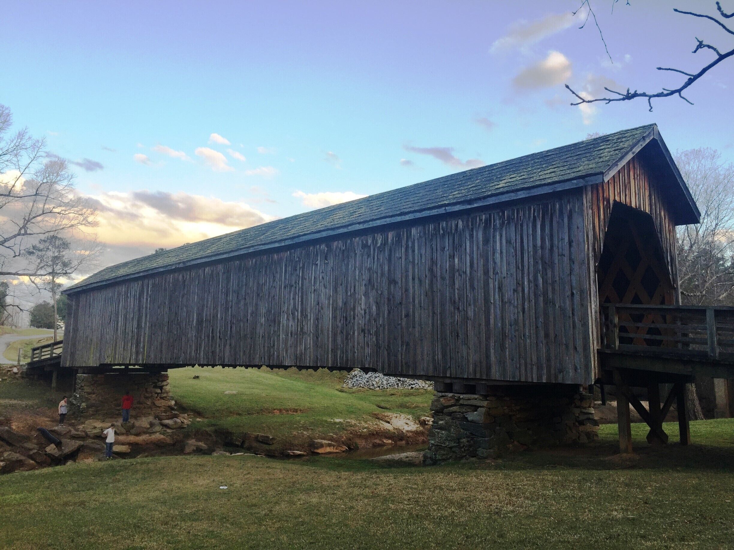 A wooden covered bridge with stone supports, crossing a shallow stream.