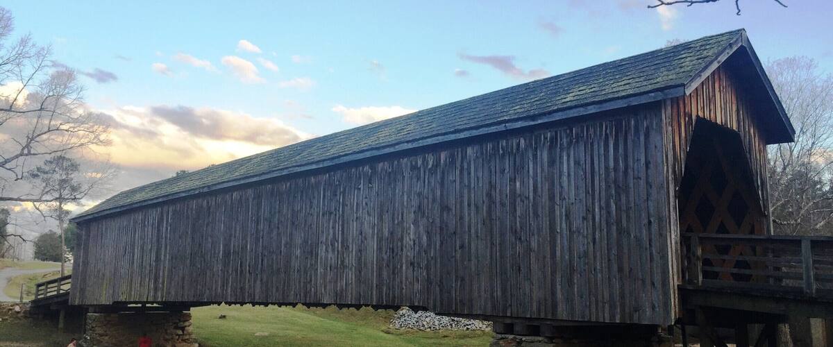 A wooden covered bridge with stone supports, crossing a shallow stream.