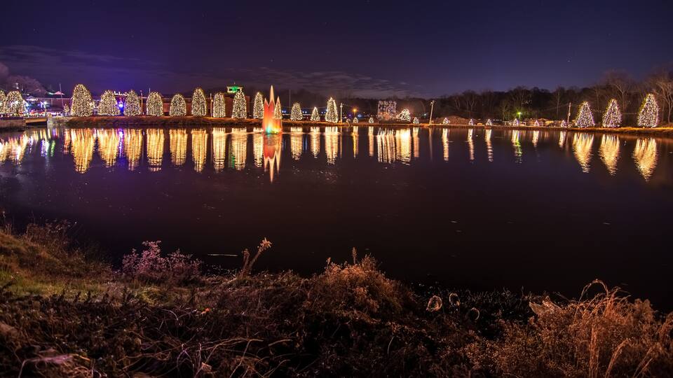 A nighttime view of a lake with illuminated Christmas trees and a fountain reflecting on the water.