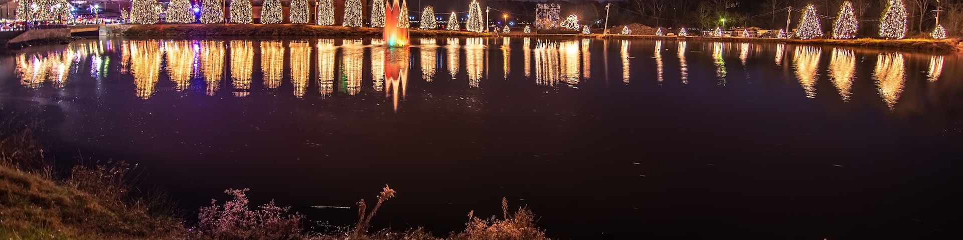 Vista noturna de um lago com árvores de Natal iluminadas e uma fonte refletindo na água.