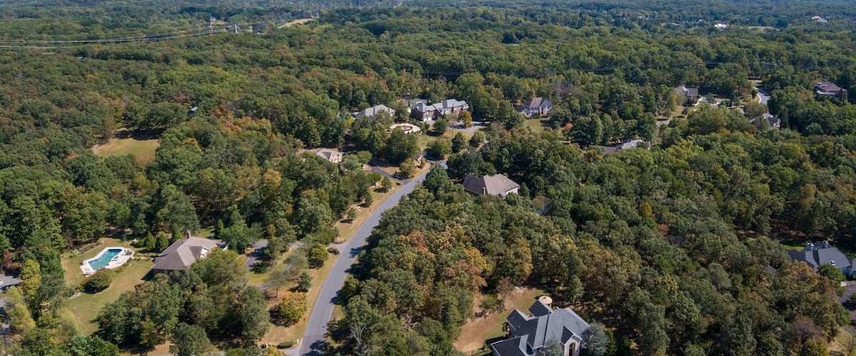 A residential area with houses surrounded by dense forest, a winding road, and a visible swimming pool.