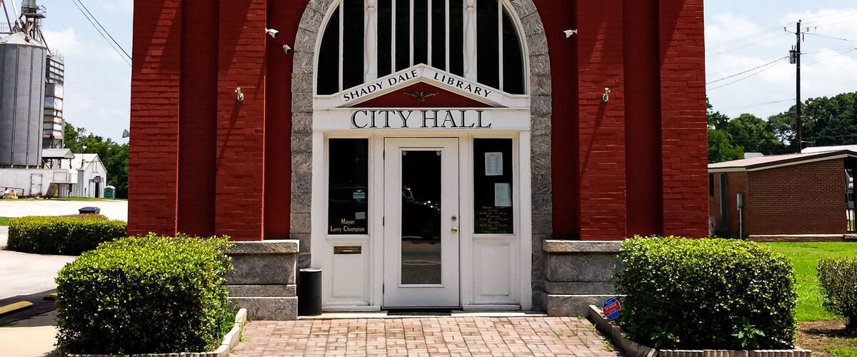 A red brick building with a white archway labeled "City Hall" and "Bank of Shady Dale Library" above it.