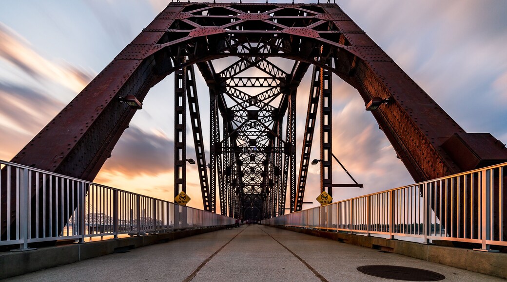 A steel bridge with a pedestrian walkway and railings, set against a sunset sky.