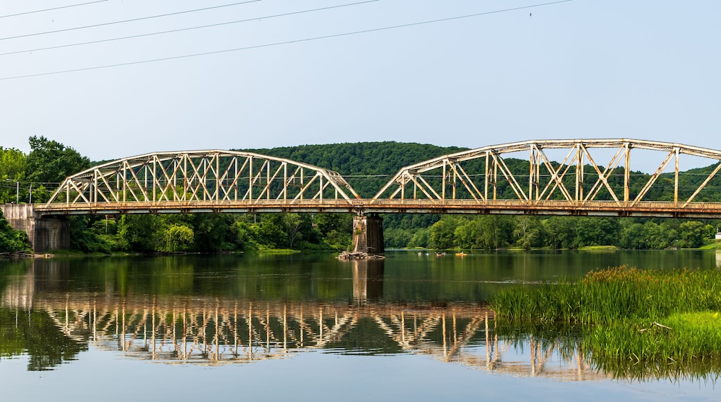 A steel truss bridge over a calm river with lush greenery and hills in the background.