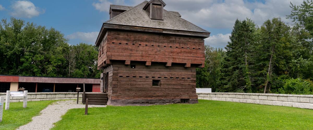 A wooden building with a sloped roof and a chimney, surrounded by a grassy area and trees.