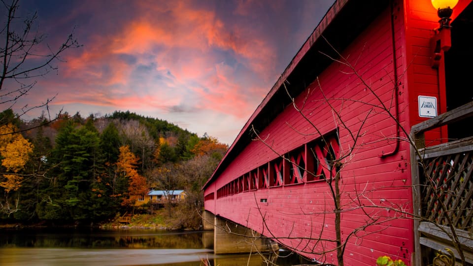 A red covered bridge over a calm river, surrounded by autumn trees and a sunset sky.