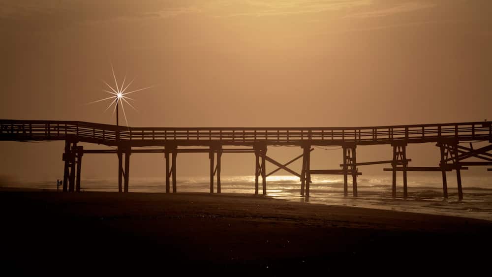Cherry Grove Fishing Pier The image shows a serene sunrise scene at North Myrtle Beach, South Carolina. A wooden pier stretches across the frame, extending out over the calm ocean waters.